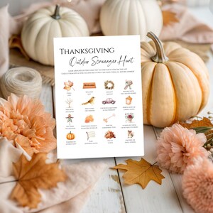 May include: A Thanksgiving-themed outdoor scavenger hunt game on a white card. The card features a list of items to find, with illustrations of a leaf, bird, car, and pumpkin. White pumpkins and orange flowers surround the card.