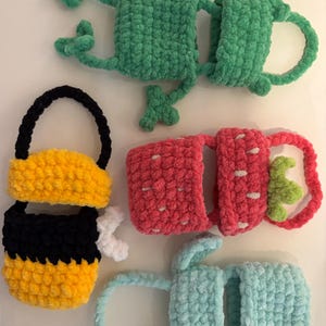 May include: A collection of handmade crochet items, including a green frog, a bee, a pink strawberry, and a light blue bunny. Each item is crafted with soft, textured yarn, featuring detailed designs and vibrant colors. The frog and bunny have handles.