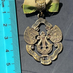Puede incluir: Una medalla de metal color latón con una lira central, dos escudos heráldicos y el texto SACHSISCHER ELBGAU SANGERBUND 1864. Está sujeta a una cinta de tela verde y crema. Una regla azul muestra que mide aproximadamente 4,5 cm de altura.
