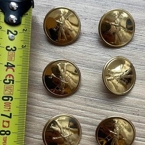 Pode incluir: Seis botões redondos de metal dourado com um design em relevo de um limpa-chaminés a carregar uma escada e uma escova. Uma fita métrica amarela está posicionada à esquerda, mostrando que os botões têm um diâmetro de cerca de 2,5 cm.