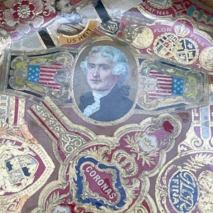 May include: A vintage cigar box lid with a colorful collage of images and text. The center features a portrait of a man with a white wig and a red, white, and blue flag. The text includes "CHAMP CLARK", "US HEIT", "RITMESTERO", "BUAT 1666", "FLOR", "FINA", "CORONAS", "ELEGANTES", and "REGALIA".