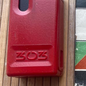 May include: A red metal lighter with the word "3023" embossed on the front. The lighter has a small rectangular opening on the top.
