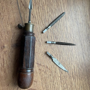 Puede incluir: Una herramienta manual antigua con mango de madera marrón oscuro, virola de latón y mecanismo de sujeción metálico. Una hoja de metal puntiaguda está insertada en el mango, mientras que otras tres hojas intercambiables están dispuestas al lado.