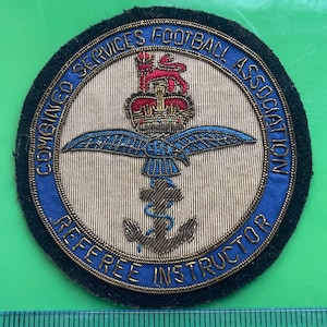 May include: A circular embroidered patch featuring the text Combined Services Football Association Referee Instructor. The design includes a red lion, a crown, blue wings, and a brown anchor on a beige background with a blue border, measuring 3 inches.