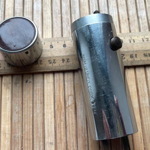 May include: A cylindrical silver-toned metal tool with a dark brown circular top and a separate tubular body featuring a small black knob and the engraved text M&M BLUHEIX DRGM. A wooden ruler is positioned behind the items for scale reference.