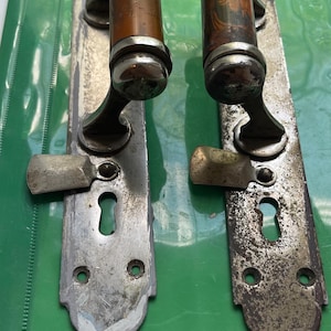 Könnte beinhalten: Zwei Vintage-Türgriffe mit langen, rechteckigen, silberfarbenen Metall-Langschildern, die Schlüssellöcher und Drehriegel aufweisen. Die Griffe haben zylindrische Griffflächen in brauner, marmorierter Holzoptik mit polierten, silberfarbenen Metallkappen.
