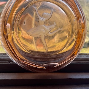 May include: A vintage pink glass ashtray with a ballerina etched into the bottom. The ballerina is in a white dress with her arms raised above her head.