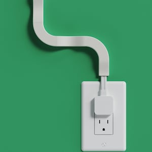 May include: A white electrical wall socket faceplate with a square plug inserted into the top outlet. A white flat cable management conduit extends upwards from the plug, curving in a serpentine shape against the solid green wall surface.