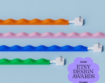 May include: Four wavy, solid-colored cable covers in orange, pink, blue, and green, each featuring a white electrical plug connector. A purple, flower-shaped badge displays the text 2026 ETSY DESIGN AWARDS Finalist in black lettering.