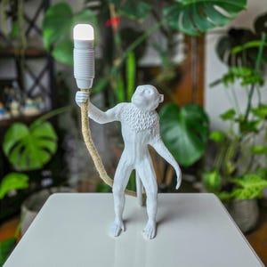 May include: White monkey lamp with a rope cord and a white light bulb. The monkey is holding the cord in its hand and is standing on a white surface.