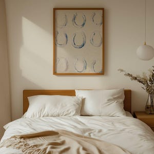 Op de afbeelding: Een slaapkamer met een houten bedframe, wit beddengoed en kussens. Een ingelijst kunstwerk met blauwe hoefijzerontwerpen hangt boven het bed. Een beige sprei voegt textuur toe. Een witte hanglamp hangt aan het plafond.