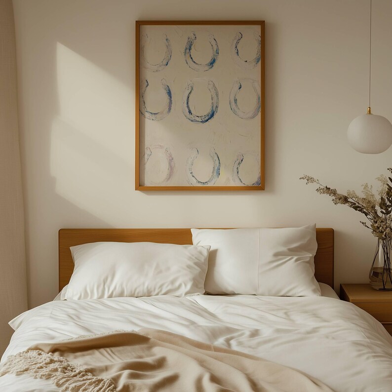 Op de afbeelding: Een slaapkamer met een houten bedframe, wit beddengoed en kussens. Een ingelijst kunstwerk met blauwe hoefijzerontwerpen hangt boven het bed. Een beige sprei voegt textuur toe. Een witte hanglamp hangt aan het plafond.