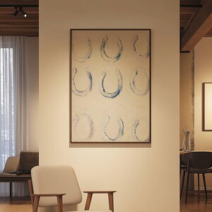 Op de afbeelding: Abstract schilderij met negen blauwe hoefijzervormen op een witte achtergrond, omlijst in donker hout. Het kunstwerk hangt aan een cr&egrave;mekleurige muur, met een neutrale stoel en bank op de voorgrond, wat een modern interieurontwerp cre&euml;ert.