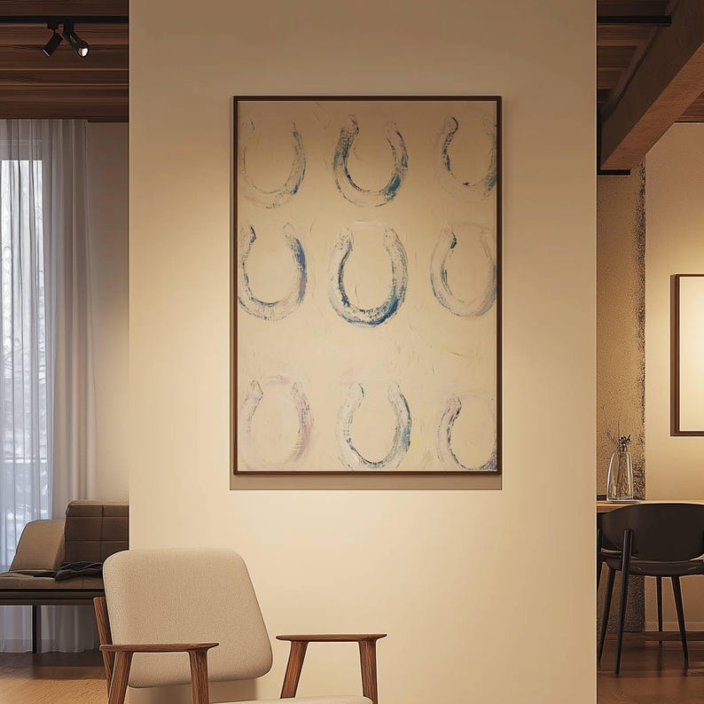 Op de afbeelding: Abstract schilderij met negen blauwe hoefijzervormen op een witte achtergrond, omlijst in donker hout. Het kunstwerk hangt aan een cr&egrave;mekleurige muur, met een neutrale stoel en bank op de voorgrond, wat een modern interieurontwerp cre&euml;ert.