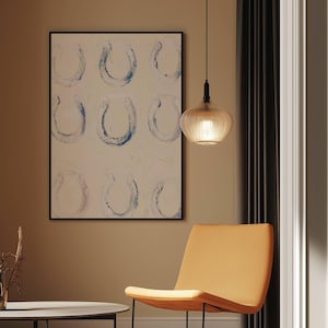 Op de afbeelding: Een abstract schilderij met negen hoefijzervormen in blauw en wit, omlijst in zwart, hangt aan een muur. Een moderne, amberkleurige hanglamp hangt aan het plafond. Een oranje stoel staat op de voorgrond.