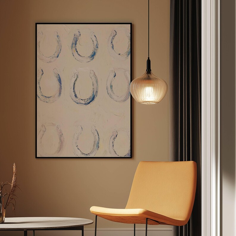 Op de afbeelding: Een abstract schilderij met negen hoefijzervormen in blauw en wit, omlijst in zwart, hangt aan een muur. Een moderne, amberkleurige hanglamp hangt aan het plafond. Een oranje stoel staat op de voorgrond.