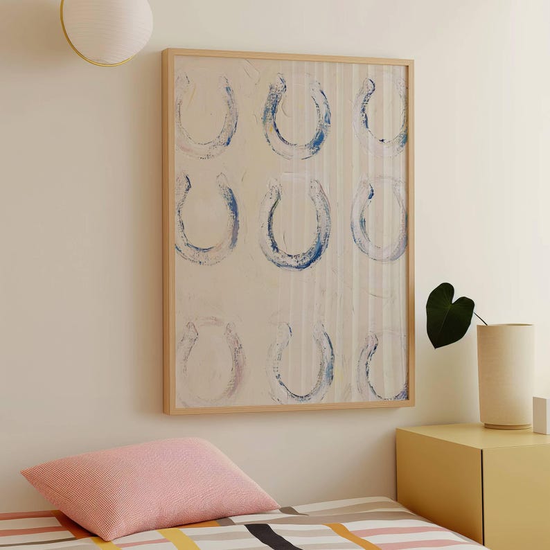 Op de afbeelding: Abstracte kunstprint met negen blauwe hoefijzervormen op een cr&egrave;mekleurige achtergrond, omlijst in licht hout. Het kunstwerk heeft een gestructureerd, verticaal gestreept patroon. Wanddecoratie.