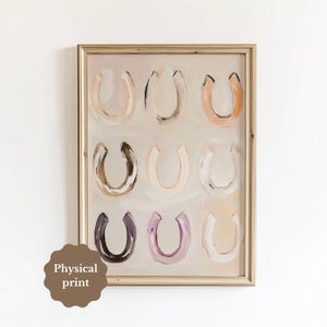 May include: A framed wall art print featuring a grid of nine painted horseshoe shapes in various neutral and muted tones including beige, brown, grey, and soft purple, set against a light cream background with a gold-coloured frame.