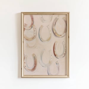 May include: A framed artwork featuring a repeating pattern of painted horseshoes in various colors, including red, yellow, and blue. The horseshoes are set against a pale pink background. The frame is a light gold color.