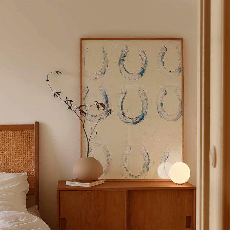 Op de afbeelding: Een houten kast met schuifdeuren bevat een beige vaas met een takje groen, een stapel boeken en een kleine, ronde, gloeiende lamp. Een groot ingelijst kunstwerk met blauwe hoefijzervormen hangt boven de kast.