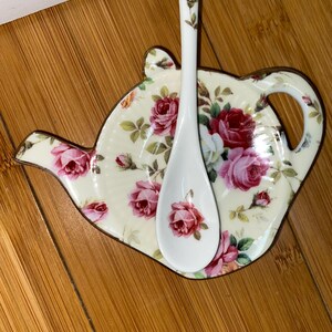 Puede incluir: Una cuchara de cerámica blanca descansa sobre un reposacucharas con forma de tetera. Ambas piezas presentan un estampado floral con rosas rosas y rojas sobre un fondo crema. El reposacucharas tiene un ribete dorado.