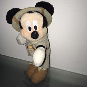 May include: A Mickey Mouse plush toy wearing a safari outfit, including a khaki-colored hat and jacket. The plush has black ears, a beige face, and brown shoes. The toy is standing on a reflective surface.