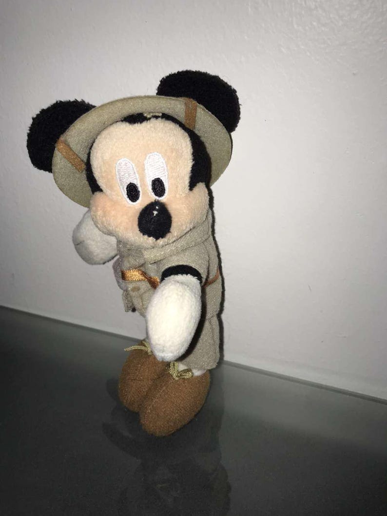 May include: A Mickey Mouse plush toy wearing a safari outfit, including a khaki-colored hat and jacket. The plush has black ears, a beige face, and brown shoes. The toy is standing on a reflective surface.