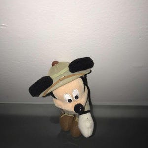 May include: A small plush toy of a cartoon mouse wearing a safari hat. The mouse has black ears, a black nose, and a white glove. The hat is tan with a brown band. The toy is tan, brown, and white.