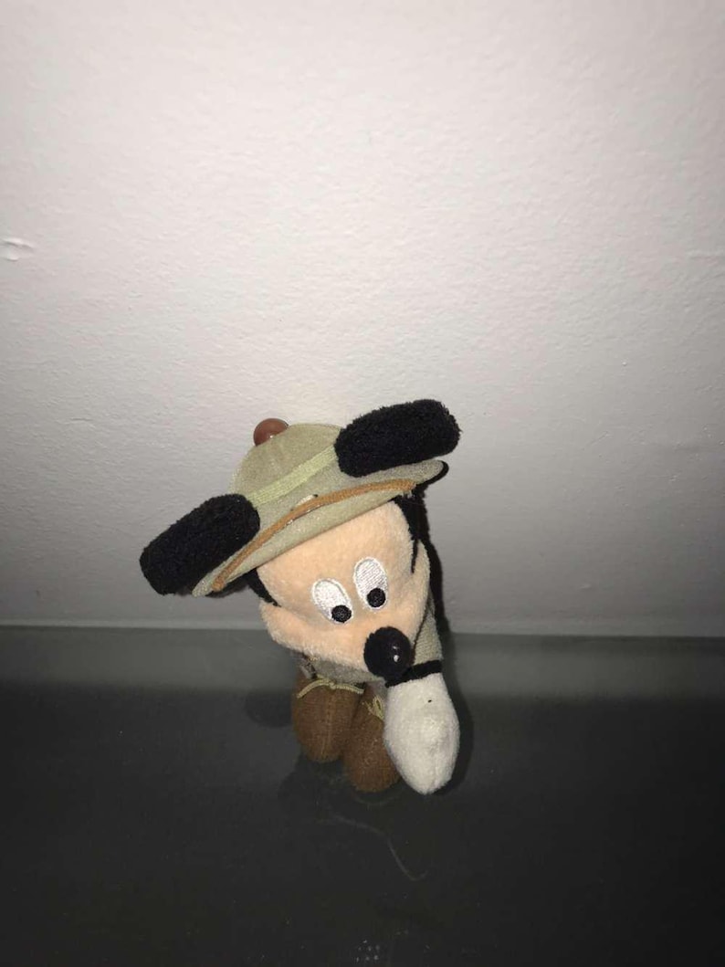 May include: A small plush toy of a cartoon mouse wearing a safari hat. The mouse has black ears, a black nose, and a white glove. The hat is tan with a brown band. The toy is tan, brown, and white.