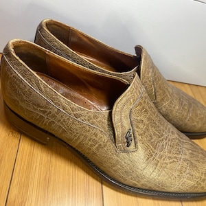 May include: A pair of brown leather slip-on shoes with a textured surface. The shoes have a classic design with a low heel and a decorative accent on the side. The shoes are made of leather and are suitable for formal or casual wear.