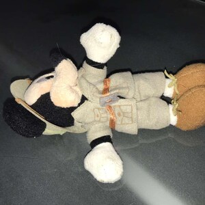 May include: A plush toy of a cartoon character, featuring a black and white head with large ears, a tan face, and a khaki outfit. The toy has brown shoes with yellow laces and white gloves.