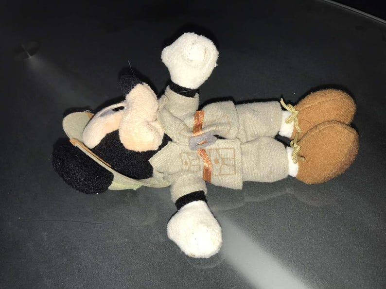 May include: A plush toy of a cartoon character, featuring a black and white head with large ears, a tan face, and a khaki outfit. The toy has brown shoes with yellow laces and white gloves.