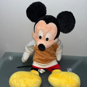 May include: A plush Mickey Mouse toy with large black ears, a tan and brown torso, and yellow feet. The toy has a black nose, white gloves, and red shorts. The Disney World tag is visible.