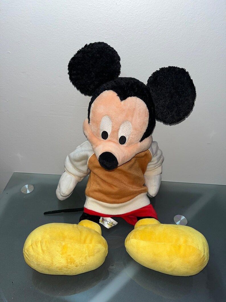 May include: A plush Mickey Mouse toy with large black ears, a tan and brown torso, and yellow feet. The toy has a black nose, white gloves, and red shorts. The Disney World tag is visible.