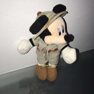 May include: A plush Mickey Mouse toy dressed in a safari outfit, including a khaki-colored hat and matching shorts and shirt. The toy has black ears, a black nose, and white gloves and is wearing brown shoes.