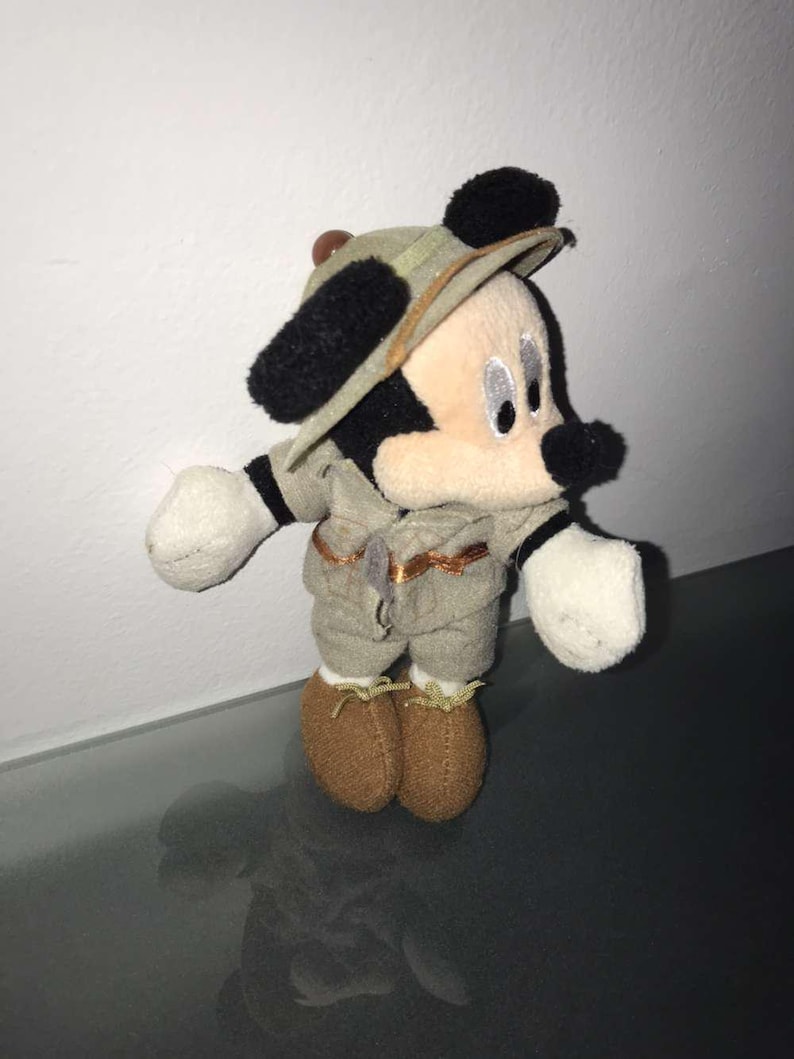 May include: A plush Mickey Mouse toy dressed in a safari outfit, including a khaki-colored hat and matching shorts and shirt. The toy has black ears, a black nose, and white gloves and is wearing brown shoes.