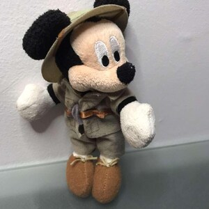May include: A plush Mickey Mouse toy dressed in a safari outfit, including a khaki-colored hat and matching jacket and pants. The toy has black ears, a black nose, and brown shoes. The toy is standing against a white background.