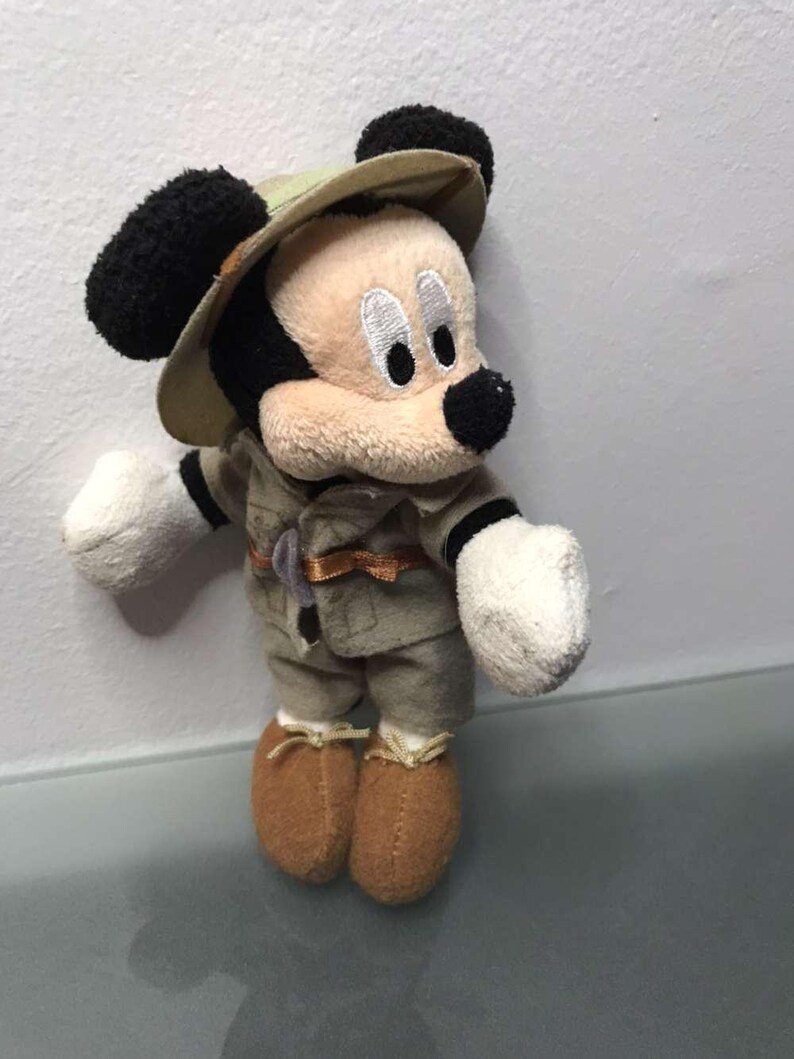 May include: A plush Mickey Mouse toy dressed in a safari outfit, including a khaki-colored hat and matching jacket and pants. The toy has black ears, a black nose, and brown shoes. The toy is standing against a white background.