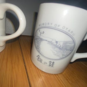 Pode incluir: Caneca de cerâmica branca com uma alça curva. A caneca apresenta uma ilustração azul e branca de uma estrada e edifícios, com o texto "MEMORY OF OTARU" e caracteres japoneses. A caneca está sobre uma superfície de madeira.
