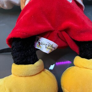 May include: A plush Mickey Mouse doll with a red velvet shorts, black legs, yellow shoes, and a brown jacket. The doll has a white tag that says "Walt Disney World".