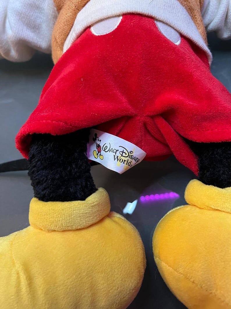 May include: A plush Mickey Mouse doll with a red velvet shorts, black legs, yellow shoes, and a brown jacket. The doll has a white tag that says "Walt Disney World".