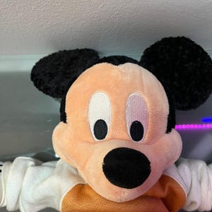 May include: A plush Mickey Mouse doll with black ears, a peach-colored face, and a black nose. The doll is wearing an orange and white sweater. The eyes are white with black pupils. The background is a light gray.