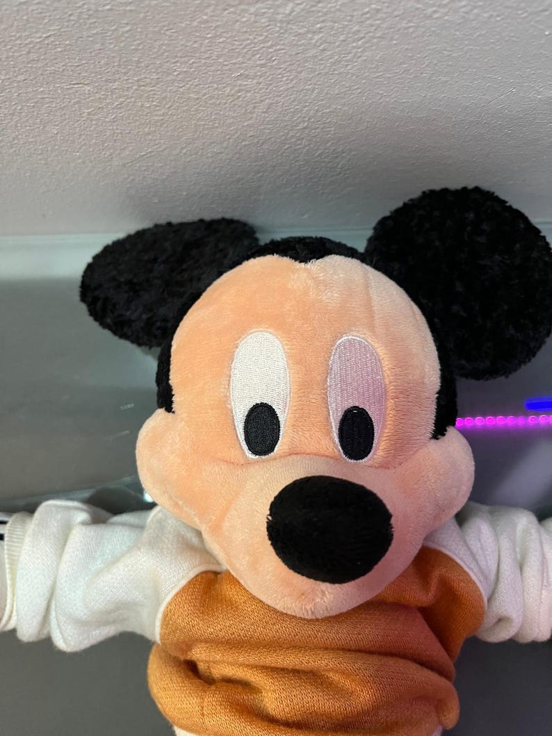 May include: A plush Mickey Mouse doll with black ears, a peach-colored face, and a black nose. The doll is wearing an orange and white sweater. The eyes are white with black pupils. The background is a light gray.