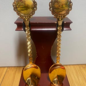 May include: A pair of ornate gold-coloured spoons, each featuring a landscape scene within a decorative frame. The handles have a twisted design. The spoons are displayed on a wooden stand, likely for decorative use.
