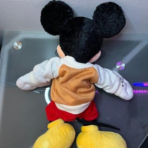 May include: A plush Mickey Mouse toy, viewed from the back. The toy features large black ears, a tan and white sweater, red shorts, and yellow shoes. The toy is lying on a reflective surface.