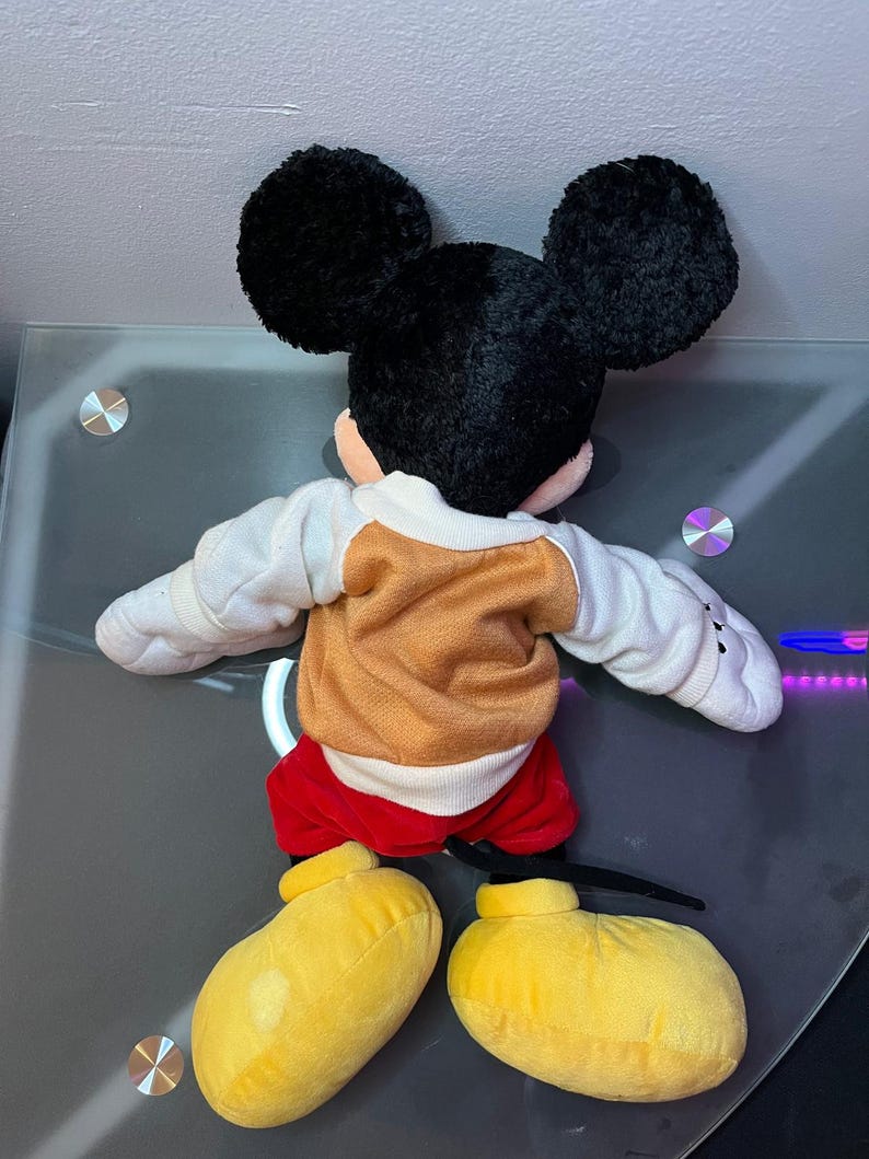 May include: A plush Mickey Mouse toy, viewed from the back. The toy features large black ears, a tan and white sweater, red shorts, and yellow shoes. The toy is lying on a reflective surface.