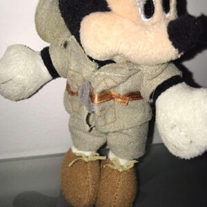 May include: A plush Mickey Mouse toy wearing a safari outfit. The toy has a black nose, ears, and eyes, with a tan face and white gloves. The outfit is khaki-colored with a brown belt and matching shoes.