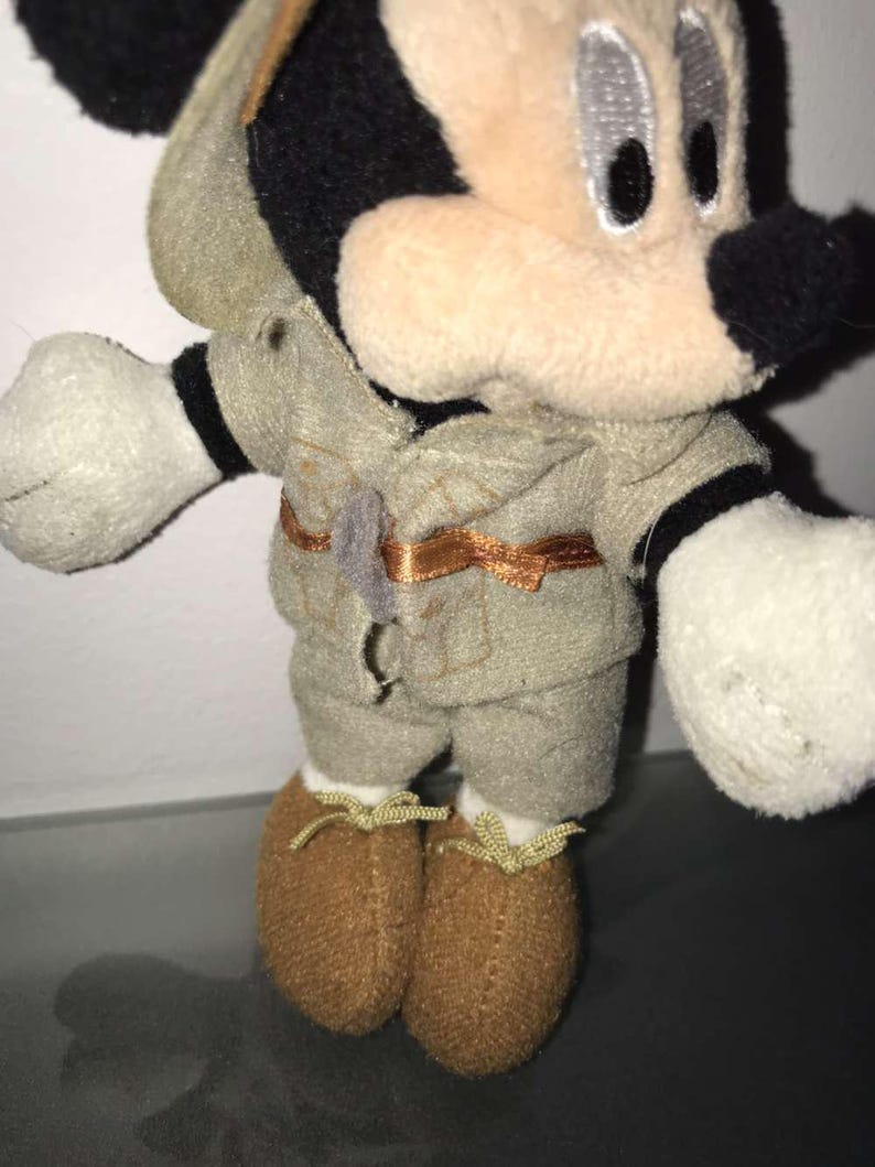 May include: A plush Mickey Mouse toy wearing a safari outfit. The toy has a black nose, ears, and eyes, with a tan face and white gloves. The outfit is khaki-colored with a brown belt and matching shoes.