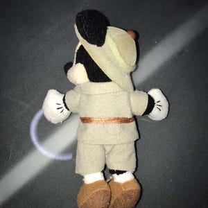 May include: A plush Mickey Mouse toy dressed in a safari outfit, including a khaki-colored hat, shirt, and shorts. The toy has black ears, a black face, white gloves with black details, and brown shoes. A brown belt accents the outfit.