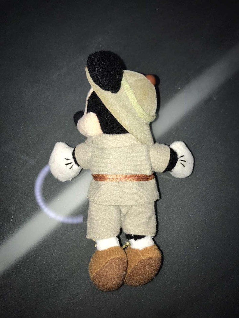 May include: A plush Mickey Mouse toy dressed in a safari outfit, including a khaki-colored hat, shirt, and shorts. The toy has black ears, a black face, white gloves with black details, and brown shoes. A brown belt accents the outfit.
